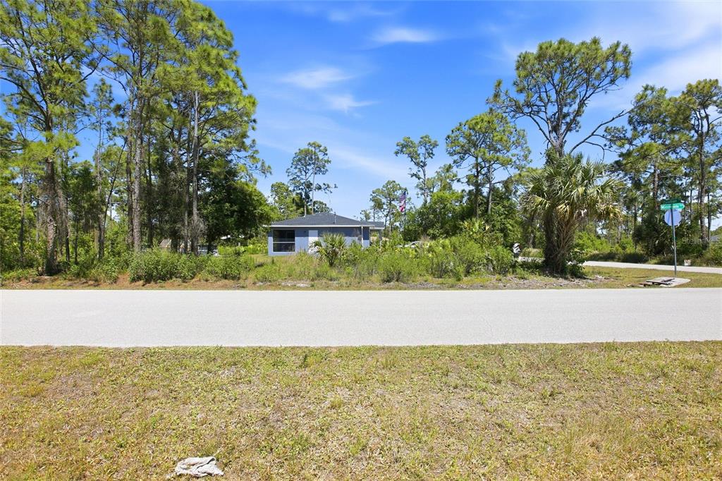 A small, blue, single-story house sits behind a paved road and dense pine trees — 27396 CORINTHIAN, DR, PUNTA GORDA, FL