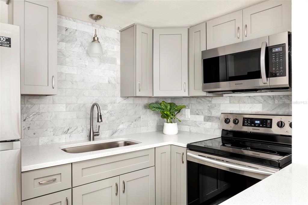 A kitchen features light gray cabinets, white countertops, a marble tile backsplash, and stainless steel appliances — 1142 SUNSET POINT ROAD, CLEARWATER, FL