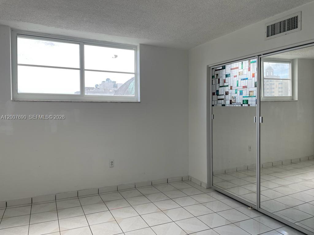 An empty room features white tiled floors, a large window, and mirrored sliding closet doors — 1750 James Ave # 7A, Miami Beach FL 33139, MIAMI BEACH, FL