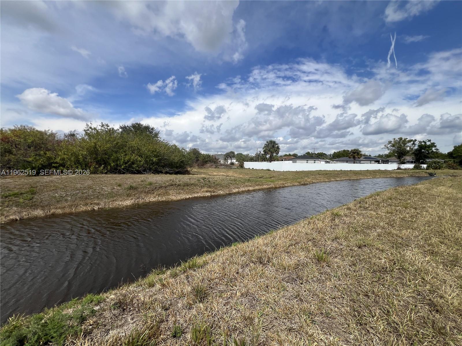 A grassy lot features a narrow canal running through the foreground under a cloudy sky — 1913 Roderick Ln, Lehigh Acres FL 33972, LEHIGH ACRES, FL