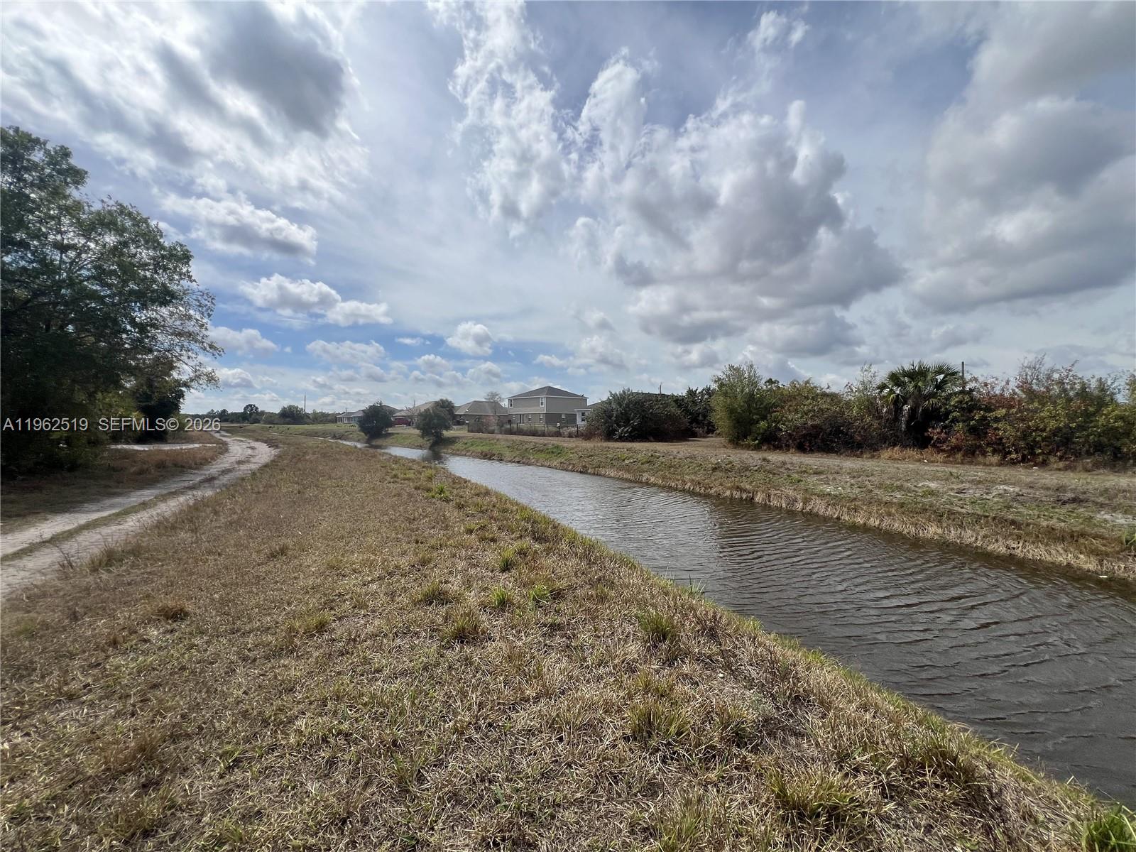 A narrow canal flows through a grassy field with houses in the distance — 1913 Roderick Ln, Lehigh Acres FL 33972, LEHIGH ACRES, FL