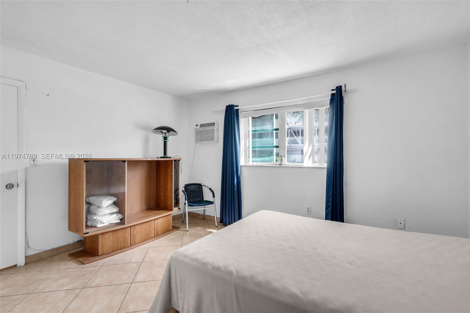 A bedroom features a bed, a wooden cabinet, a window with blue curtains, and tile flooring — 17050 NE 14th Ave # 106, Miami FL 33162, MIAMI, FL