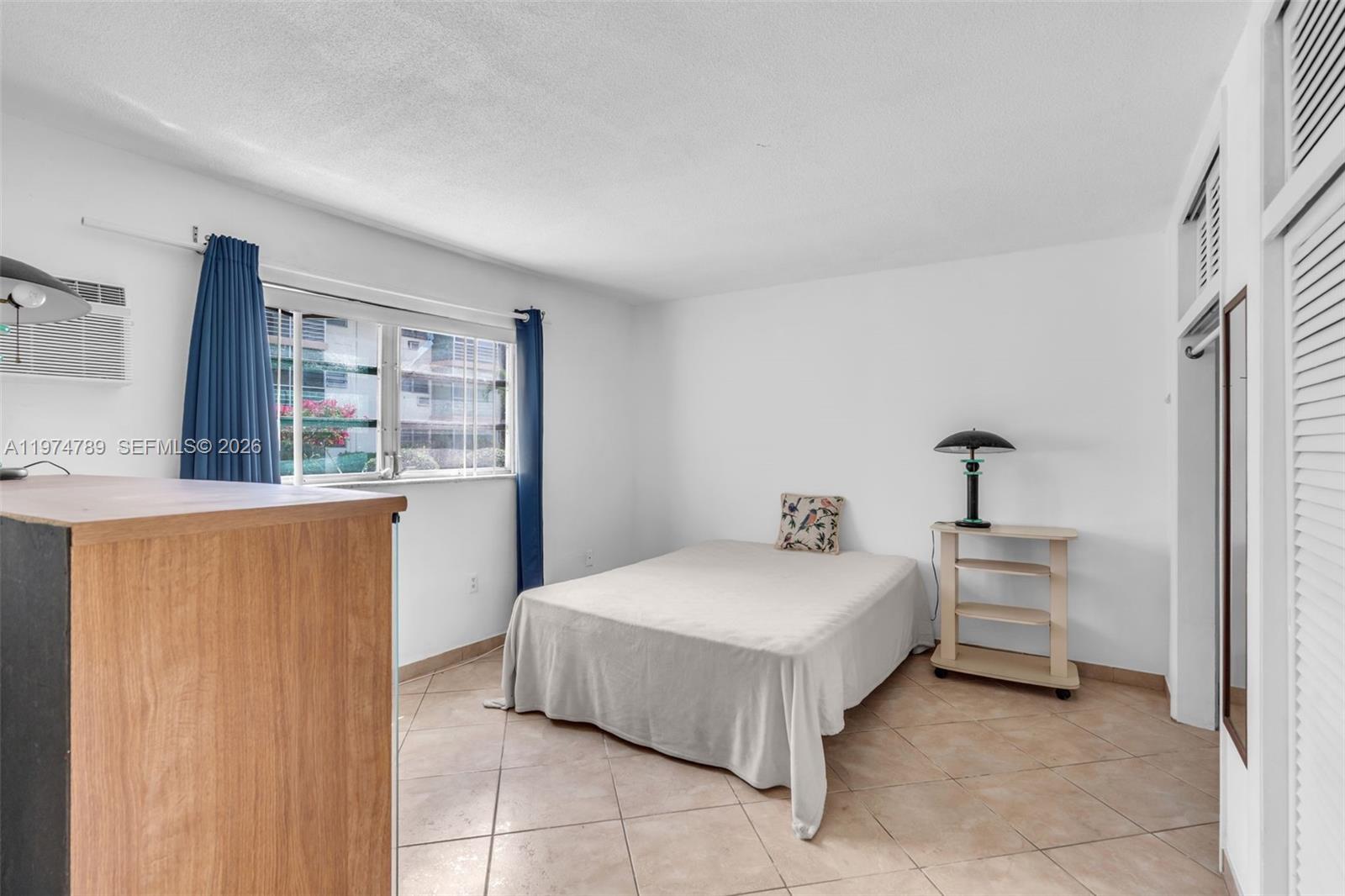 A bedroom features a bed, a small shelving unit, a window with curtains, and tile flooring — 17050 NE 14th Ave # 106, Miami FL 33162, MIAMI, FL