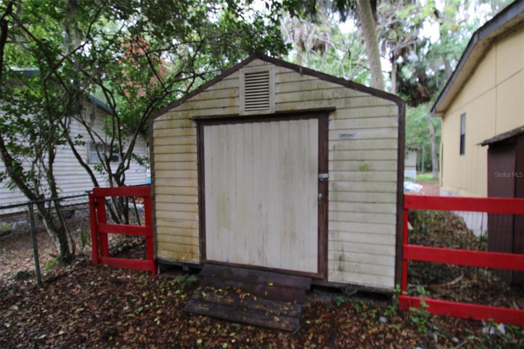 A weathered, light-colored storage shed stands between two houses behind a red wooden fence — 3128 CR 418, LAKE PANASOFFKEE, FL