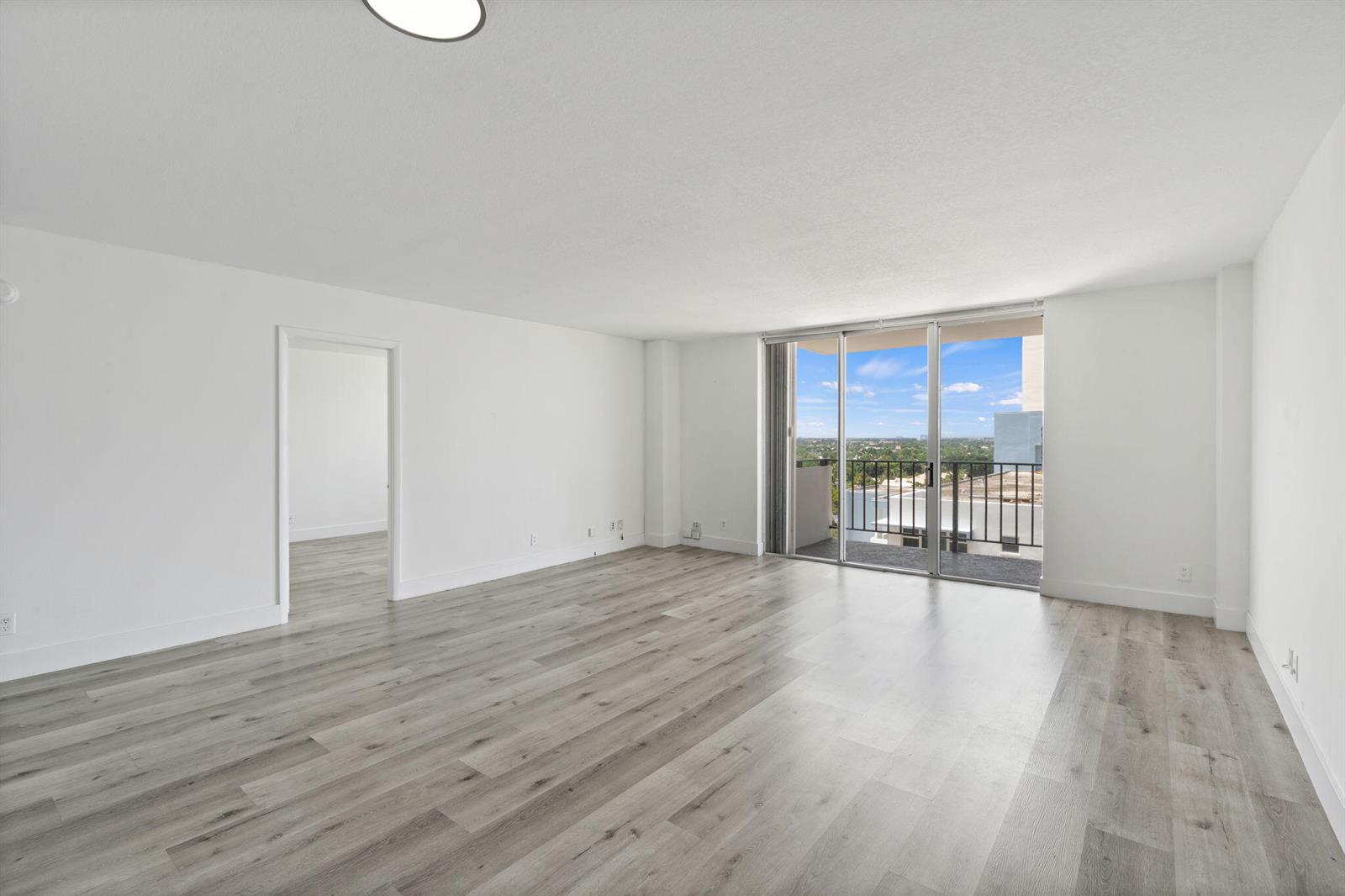 An empty living room features light wood-look flooring, white walls, and a balcony with views — 1900 S Ocean Boulevard # 10j, Pompano Beach FL 33062, POMPANO BEACH, FL