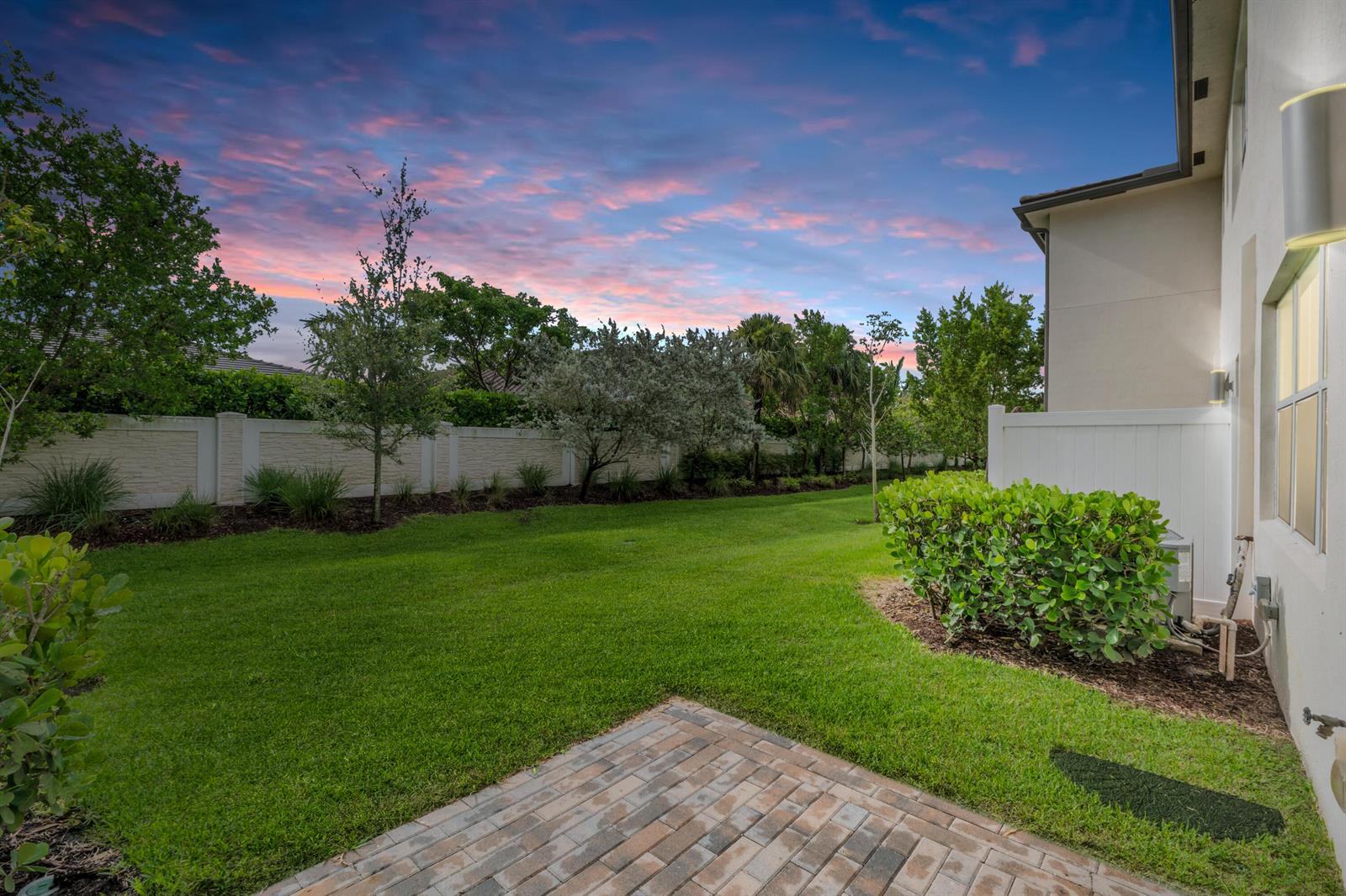 A grassy backyard features a brick patio, a white privacy fence, and mature landscaping at sunset — 4969 Navali Drive, Lake Worth FL 33467, LAKE WORTH, FL