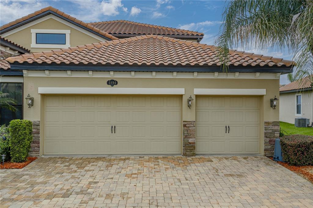 A beige house exterior features a three-car garage with a paver driveway and tile roof — 1894 VIA LAGO DRIVE, LAKELAND, FL