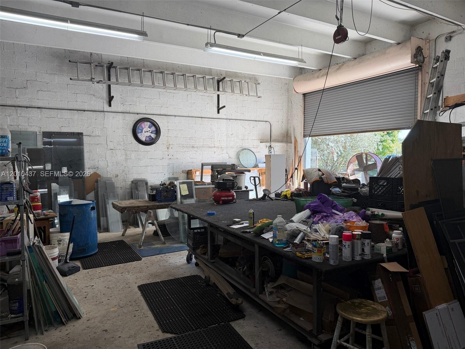 A cluttered workshop features a large central workbench, concrete block walls, and a roll-up door — 10 NE 3rd St, Florida City FL 33034, FLORIDA CITY, FL