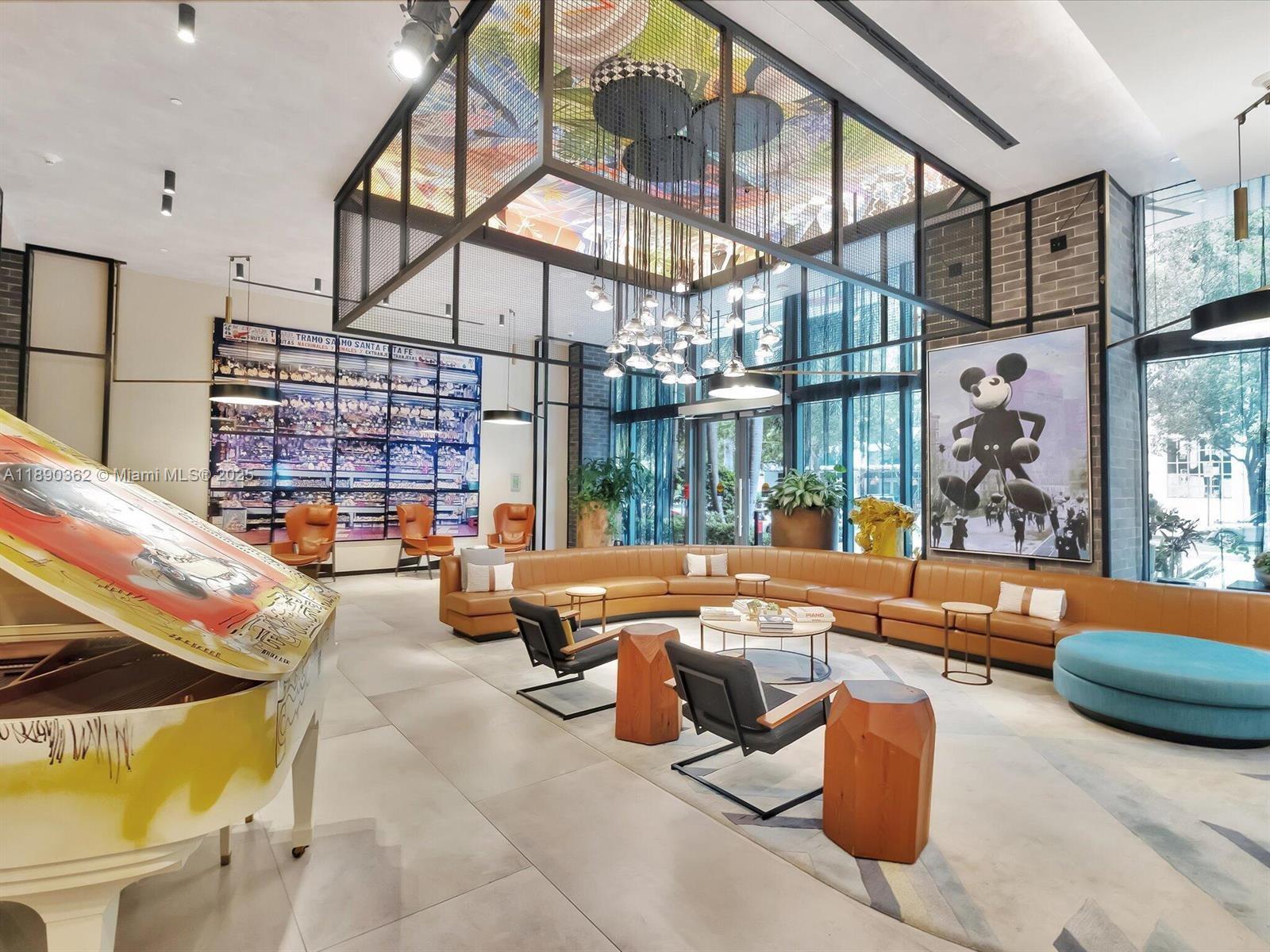 A modern lobby features a grand piano, a large curved sofa, and a Mickey Mouse painting — 121 NE 34th St # 1610, Miami FL 33137, MIAMI, FL