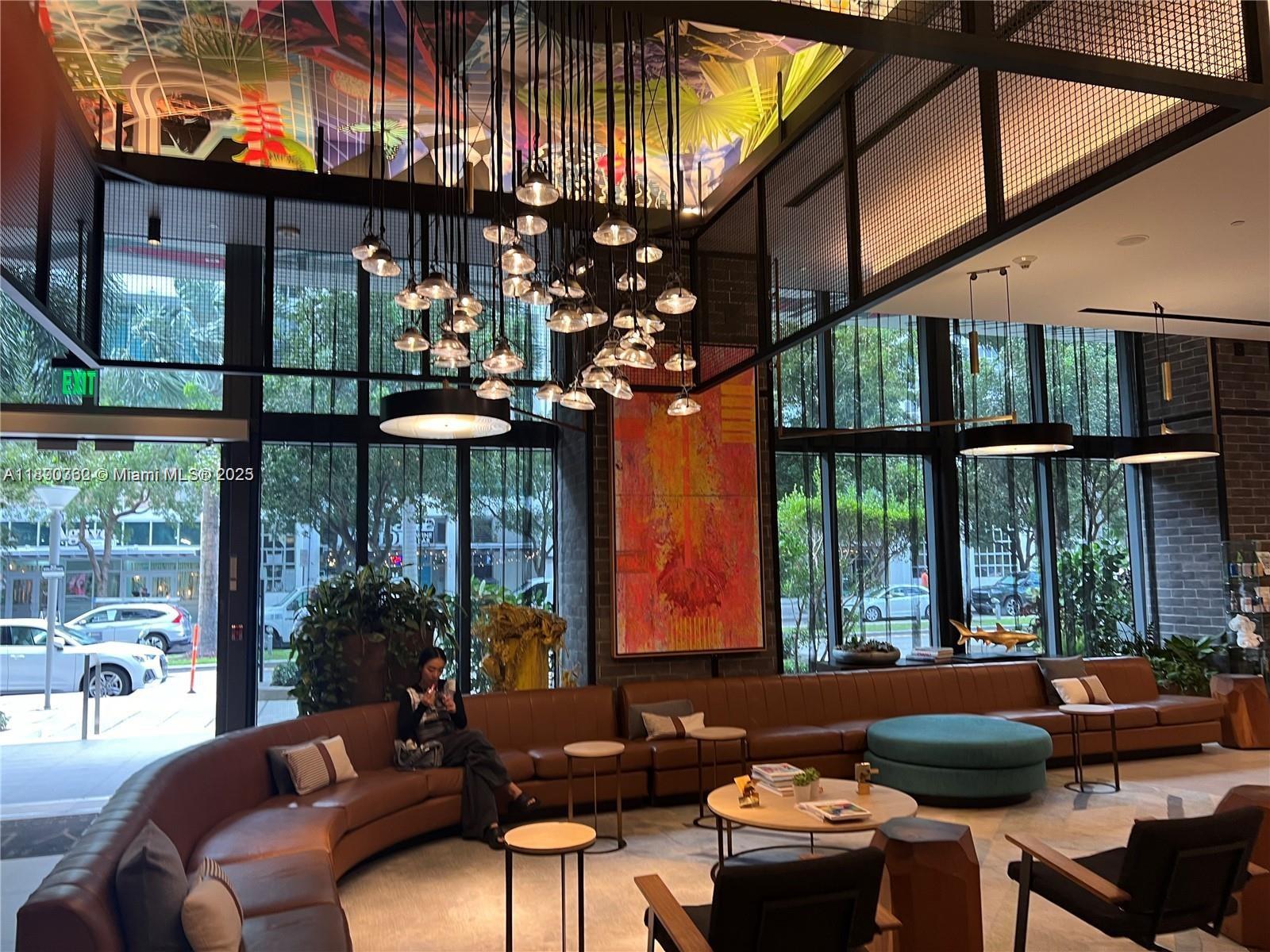 A modern lobby features a curved leather sofa, a large chandelier, and floor-to-ceiling windows — 121 NE 34th St # 1610, Miami FL 33137, MIAMI, FL