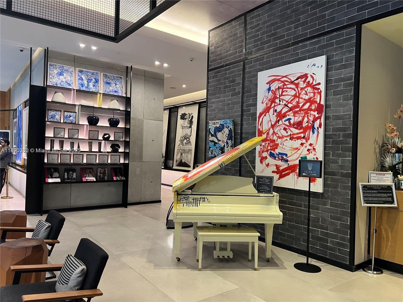 A modern lobby features a white grand piano, abstract wall art, and built-in shelving — 121 NE 34th St # 1610, Miami FL 33137, MIAMI, FL