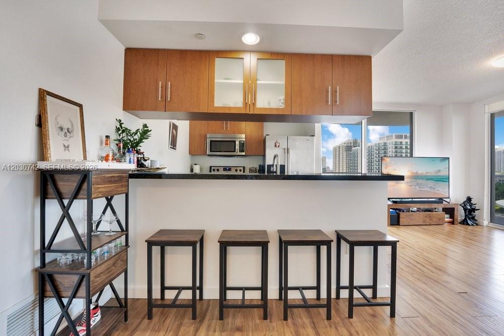 A kitchen with wooden cabinets and a breakfast bar featuring four stools faces a living area — 601 NE 23rd St # 1406, Miami FL 33137, MIAMI, FL