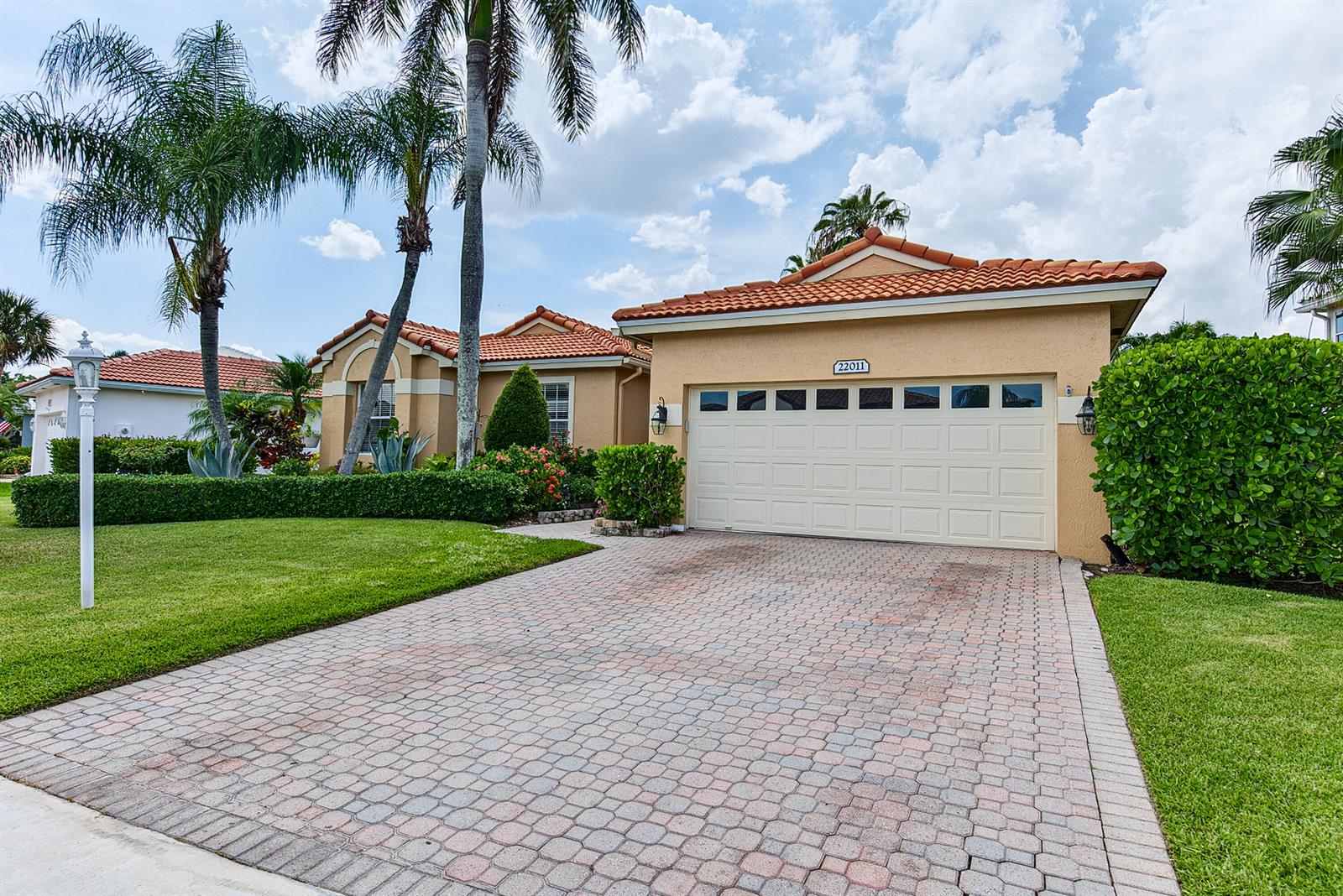A tan house with a tile roof and a paved driveway sits behind a manicured hedge — 22011 Palm Grass Drive, Boca Raton FL 33428, BOCA RATON, FL