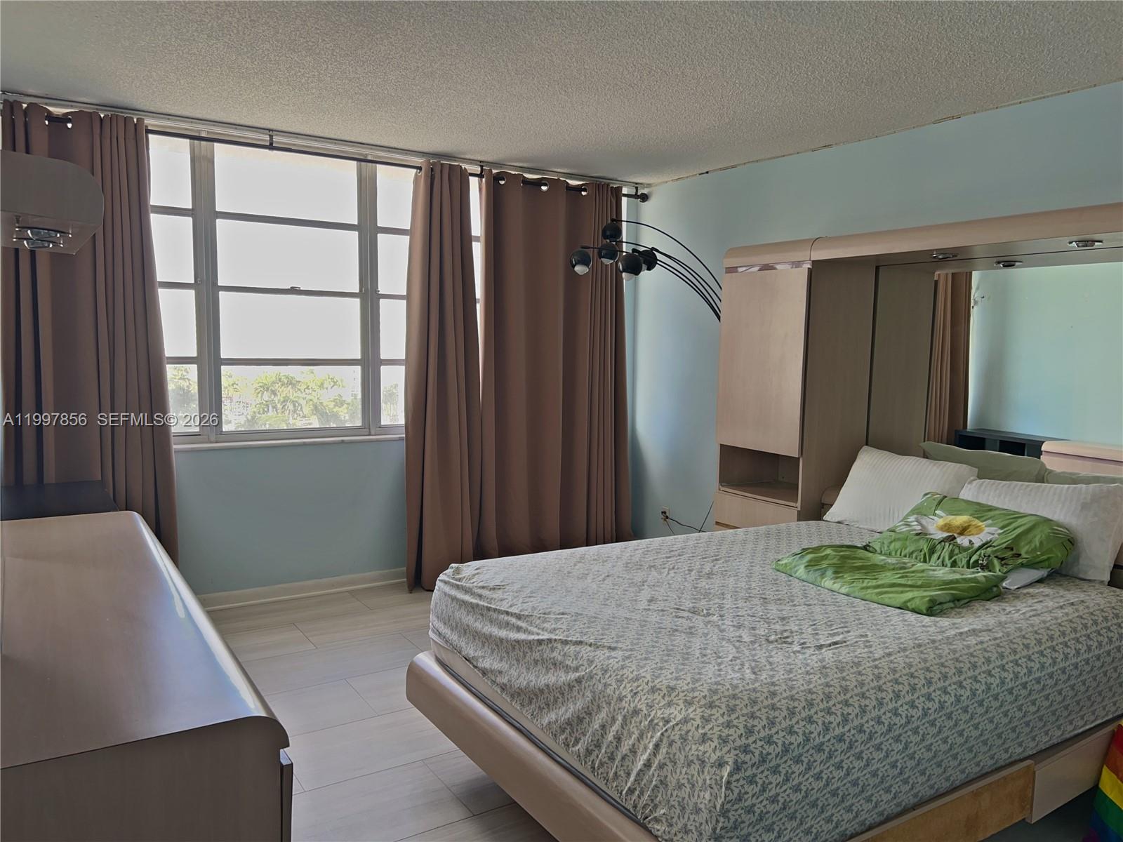 A bedroom features light blue walls, a large window with brown curtains, and a bed — 1801 S Ocean Dr # 934, Hallandale Beach FL 33009, HALLANDALE BEACH, FL
