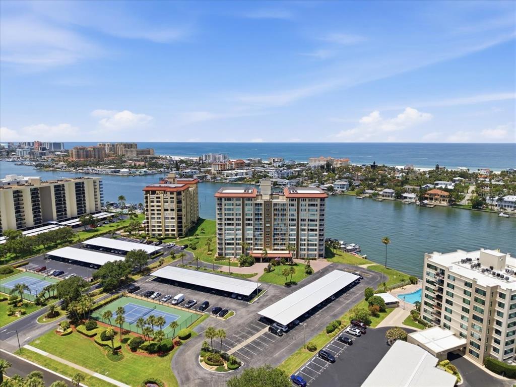An aerial view shows multiple high-rise apartment buildings, parking lots, and tennis courts near water — 736 ISLAND WAY UNIT 1003, CLEARWATER BEACH, FL
