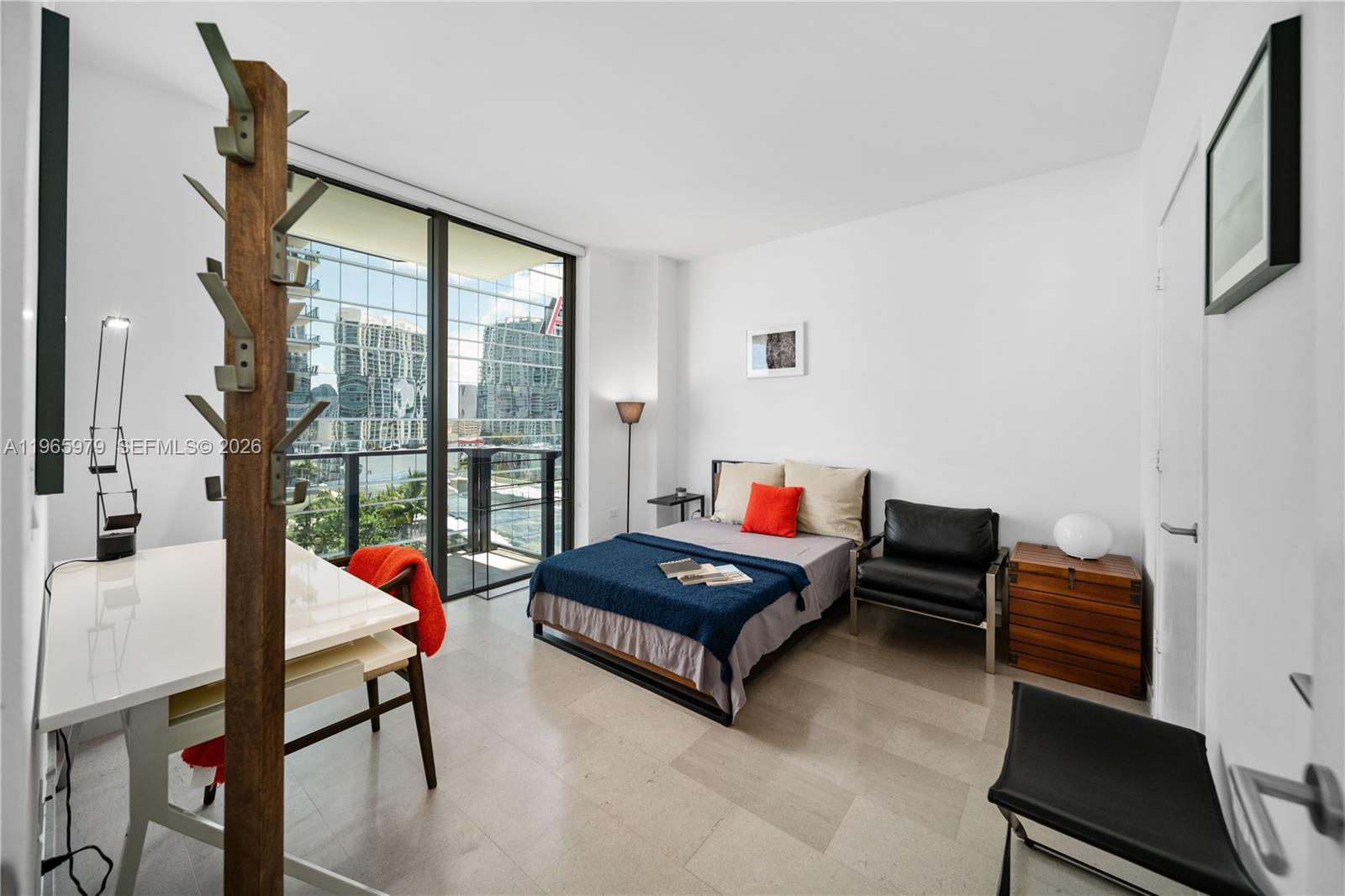 A modern bedroom features a bed, desk, and floor-to-ceiling windows overlooking a city skyline — 68 SE 6th St # 1201, Miami FL 33131, MIAMI, FL