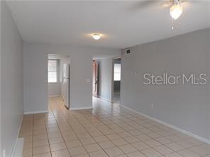 A room with light gray walls and beige tiled flooring leads to a kitchen area — 2929 W OAK RIDGE ROAD UNIT E8, ORLANDO, FL