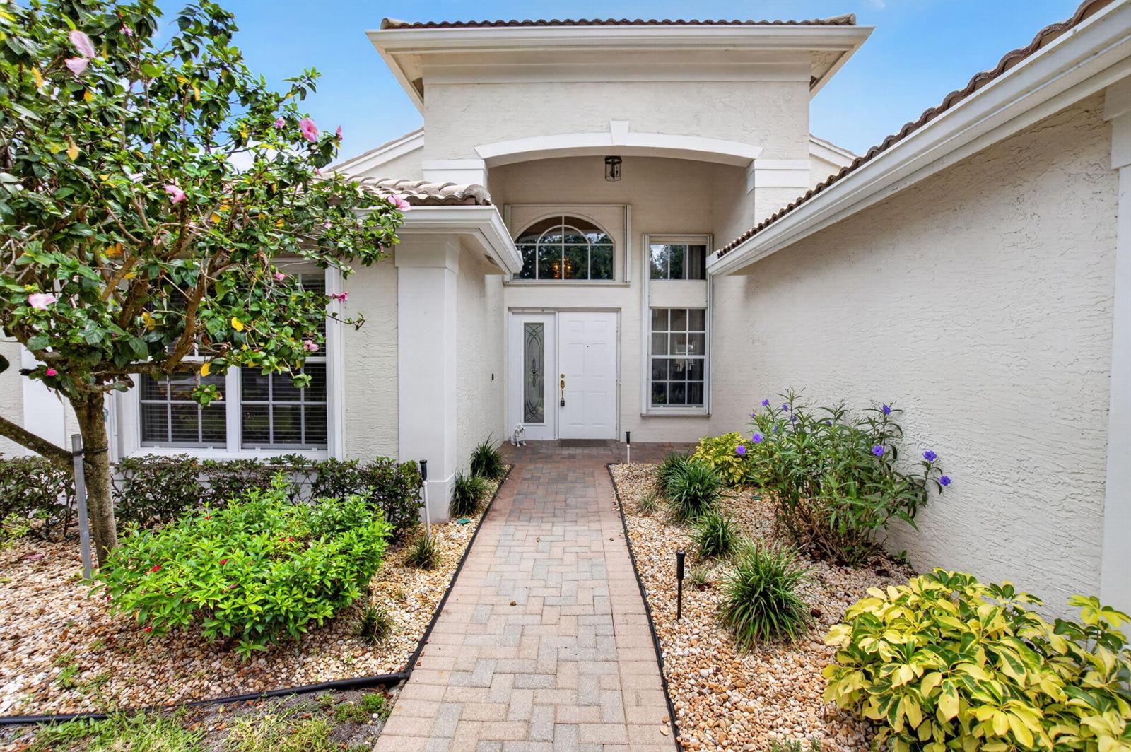 A paved walkway leads to the white front door of a stucco house with landscaping — 13461 Barcelona Lake Circle, Delray Beach FL 33446, DELRAY BEACH, FL