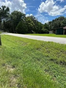 A grassy lot sits beside a paved road with trees and a small building nearby — 26408 ROLLING ACRES DRIVE, BROOKSVILLE, FL