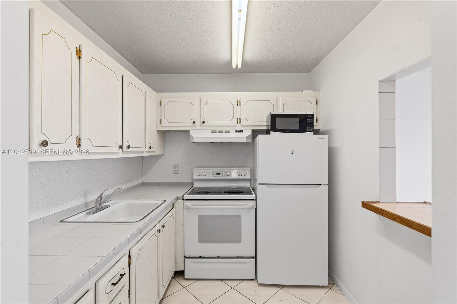 A small kitchen features white cabinets, white appliances, tiled countertops, and a pass-through window — 237 Camden J # 237, West Palm Beach FL 33417, WEST PALM BEACH, FL