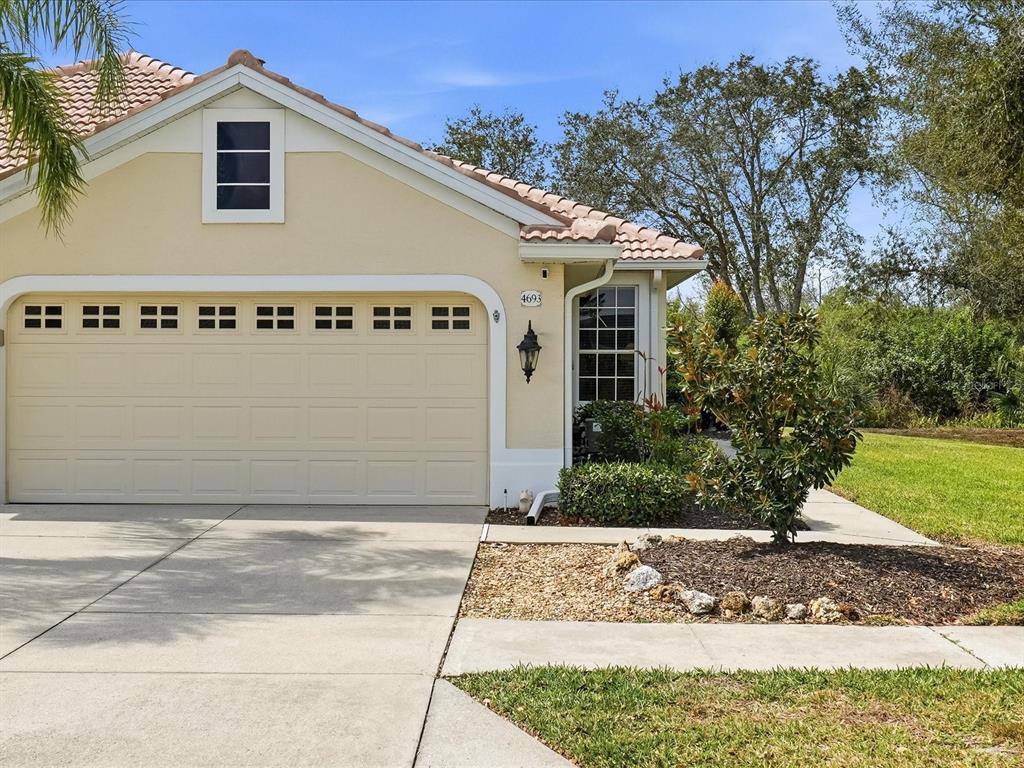 A beige house exterior features a two-car garage, a tiled roof, and a small garden — 4693 WHISPERING OAKS DRIVE, NORTH PORT, FL