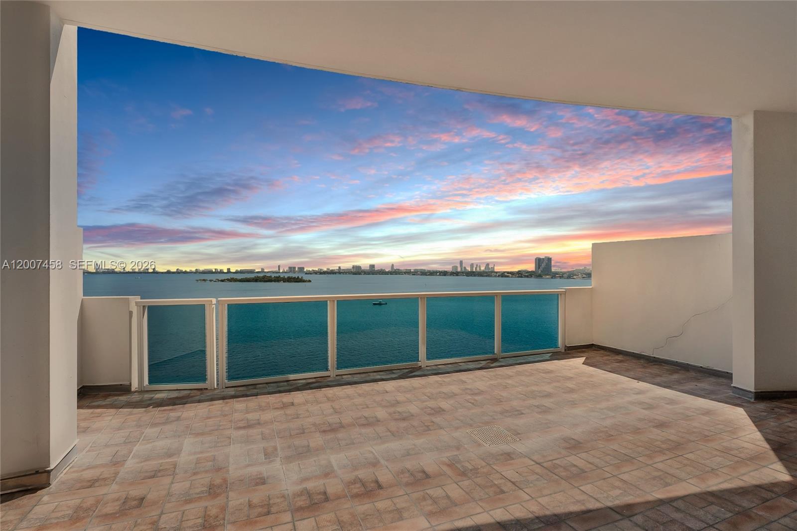 A spacious balcony features tiled flooring and a glass railing overlooking a bay at sunset — 725 NE 22nd St # 7B, Miami FL 33137, MIAMI, FL