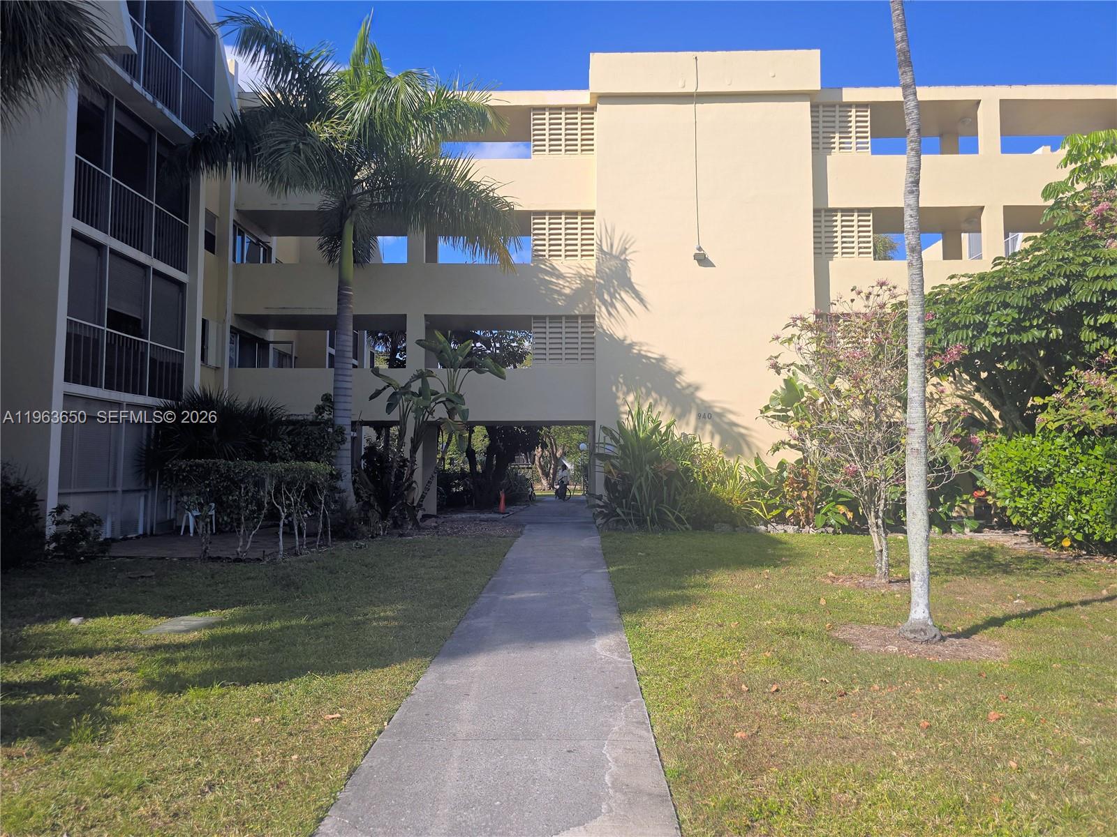 A concrete walkway leads to the entrance of a multi-story tan apartment building — 940 NE 199th St # 114, Miami FL 33179, MIAMI, FL
