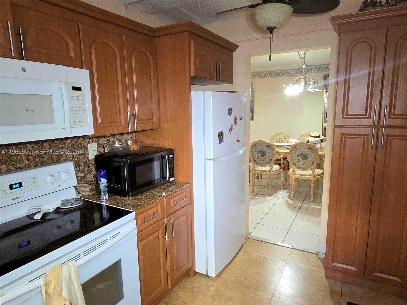 A kitchen with wood cabinets, white appliances, and granite countertops leads into a dining room — 940 NE 199th St # 114, Miami FL 33179, MIAMI, FL