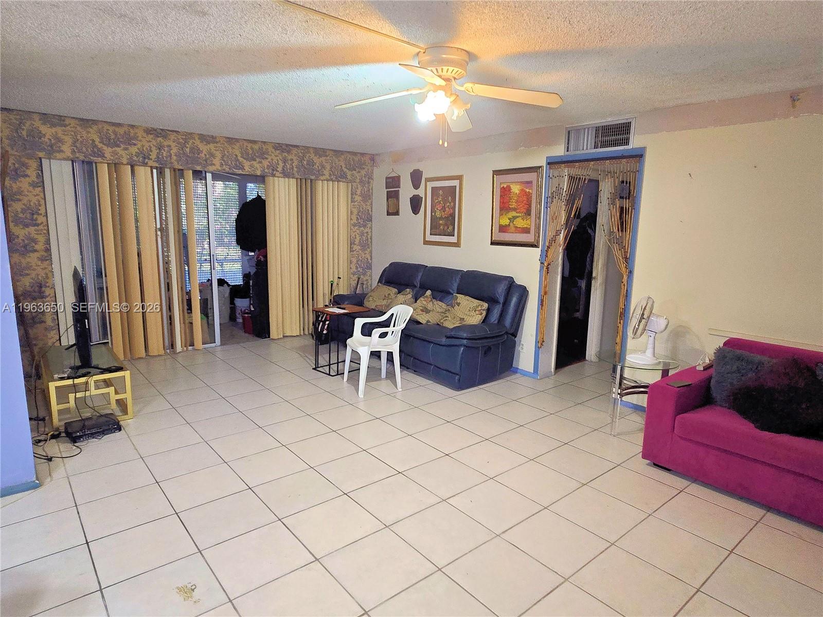 A living room features white tile flooring, a blue sofa, a magenta chair, and a ceiling fan — 940 NE 199th St # 114, Miami FL 33179, MIAMI, FL