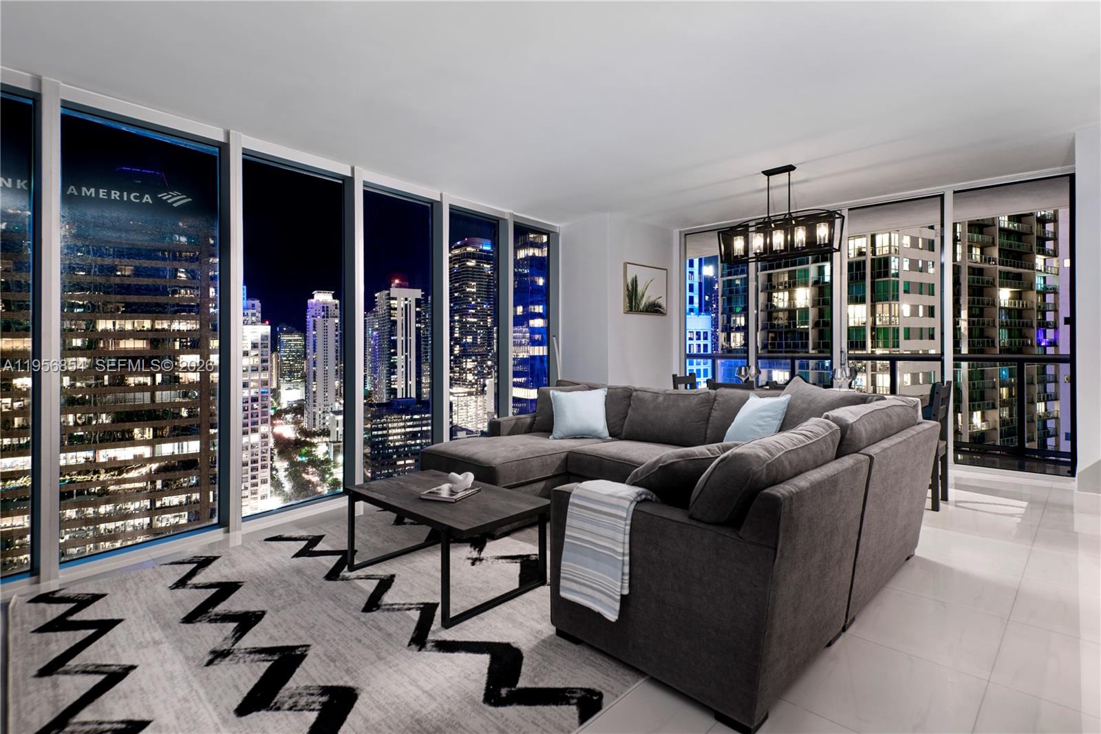 A modern living room features a gray sectional sofa, patterned rug, and floor-to-ceiling city views — 485 Brickell Ave # 3208, Miami FL 33131, MIAMI, FL