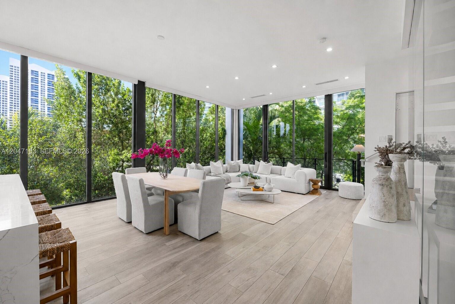 A modern, open-concept living and dining area features floor-to-ceiling windows overlooking lush green trees — 1525 Bay Rd # 1525, Miami Beach FL 33139, MIAMI BEACH, FL