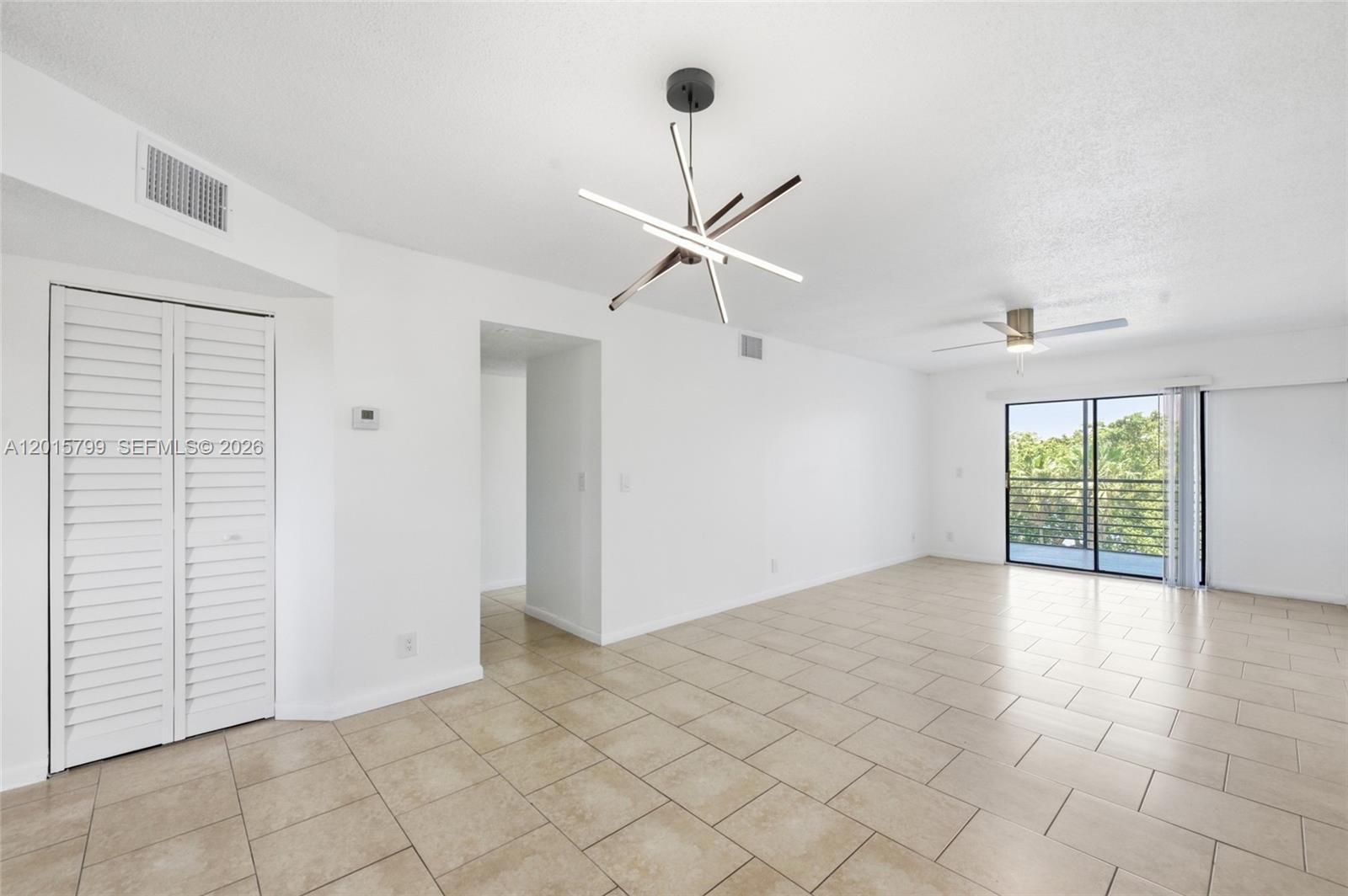 An empty living room features white walls, beige tile flooring, a modern chandelier, and balcony access — SUNRISE, FL