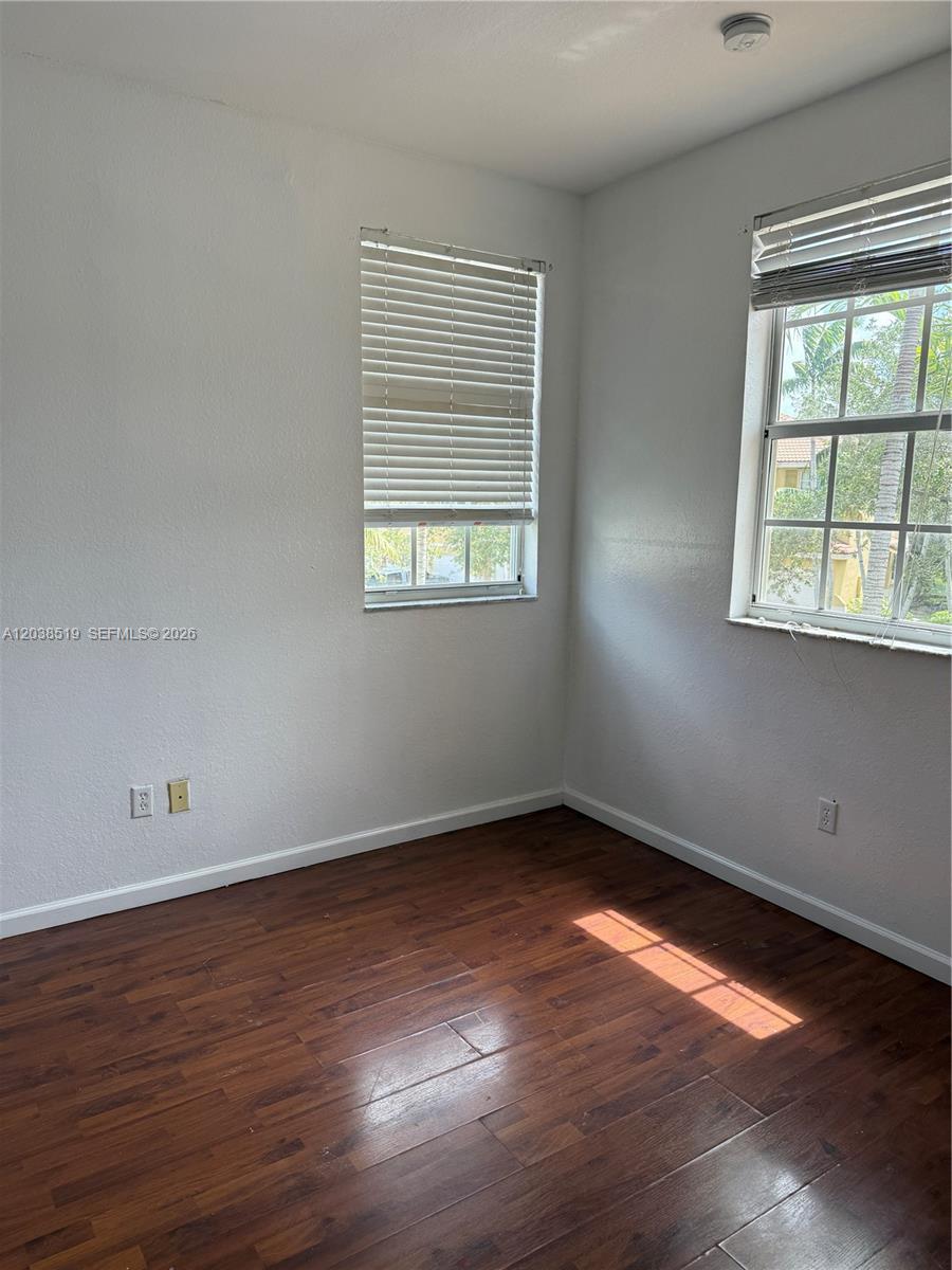 An empty room features dark wood-look flooring, white walls, and two windows with blinds — 551 NE 21st Ter, Homestead FL 33033, HOMESTEAD, FL