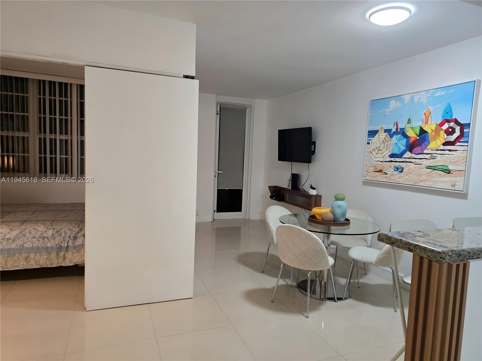 A dining area with a glass table and white chairs sits beside a bedroom partition — 100 Lincoln Rd # 503, Miami Beach FL 33139, MIAMI BEACH, FL