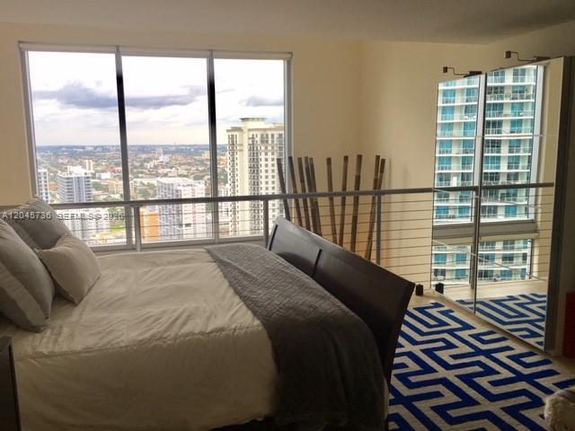 A bedroom features a bed, a blue patterned rug, and floor-to-ceiling windows overlooking a city — MIAMI, FL