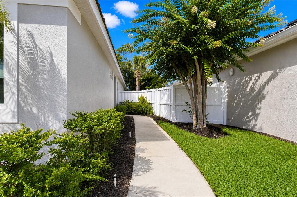 A concrete walkway leads between two light-colored house walls toward a white privacy fence — 8644 54TH AVENUE CIRCLE E, BRADENTON, FL