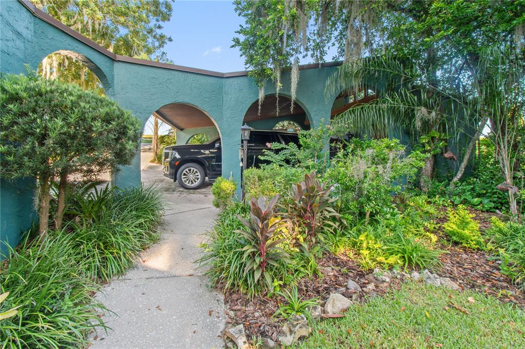 A teal stucco carport with arched openings sits beside a landscaped garden and walkway — 1019 W LAKE ELOISE TERRACE, WINTER HAVEN, FL