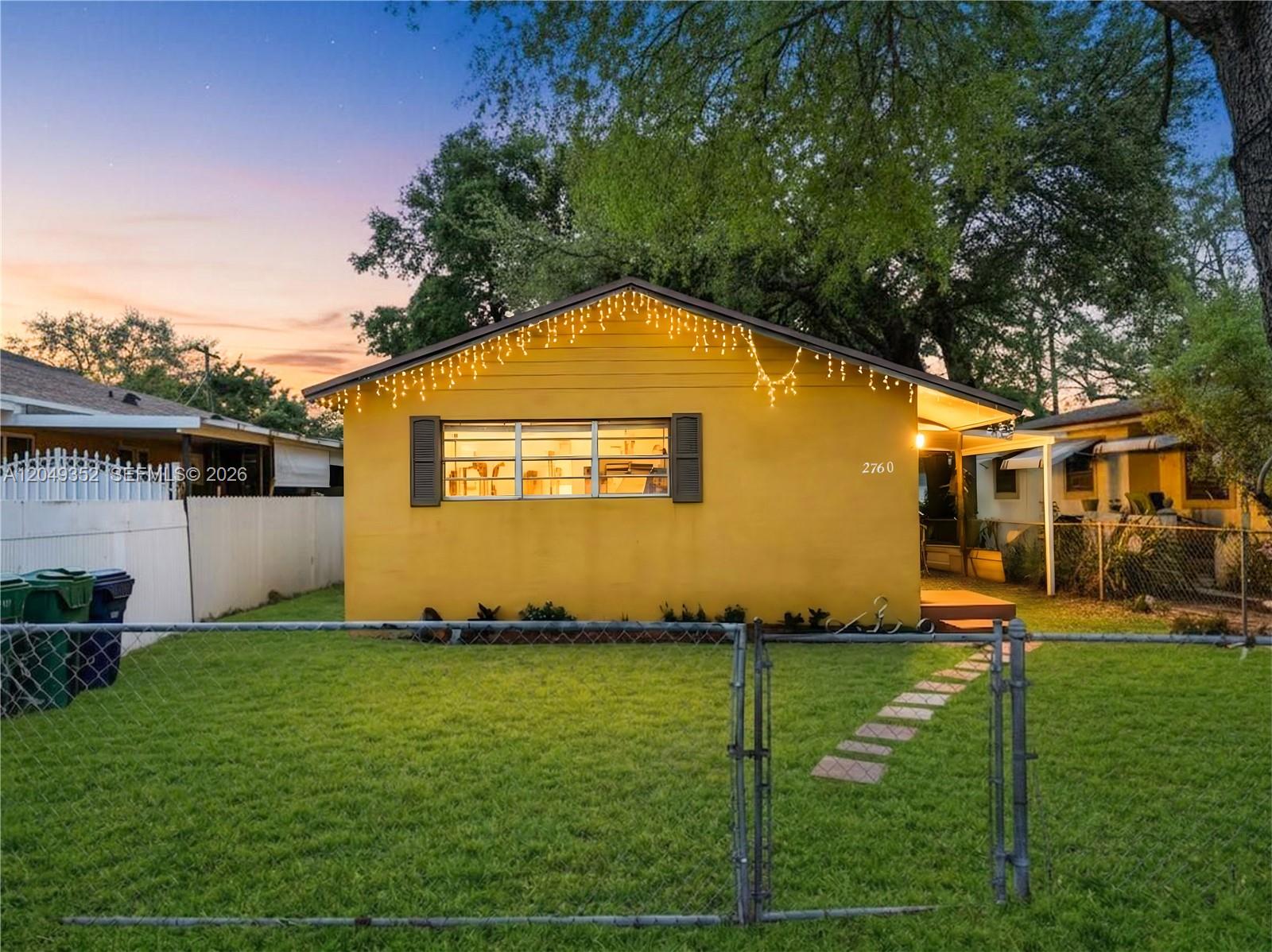 A yellow single-story house with a chain-link fence and string lights at dusk — 2760 NW 58th St, Miami FL 33142, MIAMI, FL