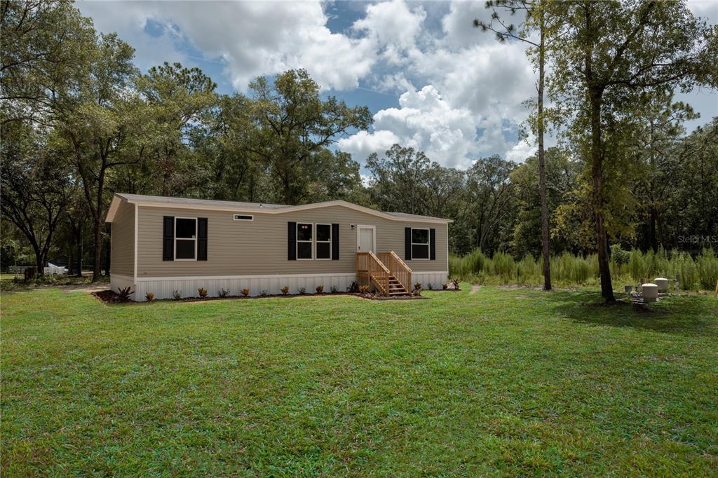 A single-story manufactured home with light tan siding sits on a grassy, wooded lot — 104 JACKSON AVENUE, INTERLACHEN, FL