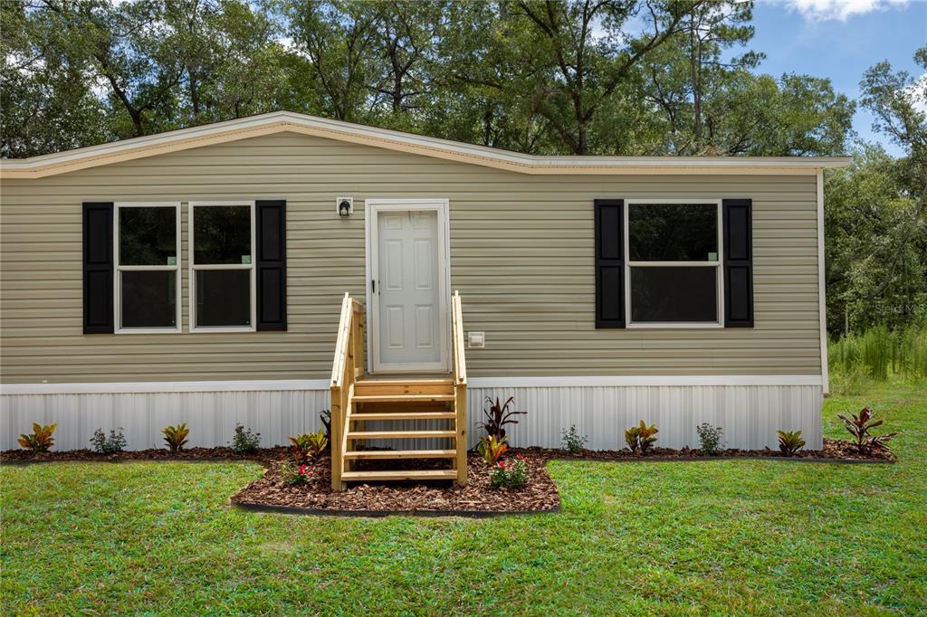 A tan manufactured home with black shutters and wooden stairs leading to the front door — 104 JACKSON AVENUE, INTERLACHEN, FL
