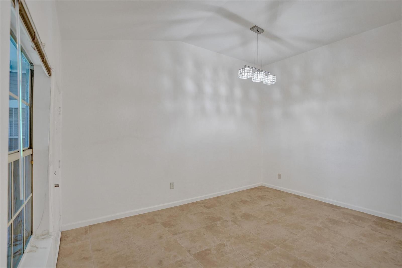 An empty room features white walls, tan tile flooring, and a modern three-light crystal chandelier — 1050 Crystal Way, Delray Beach FL 33444, DELRAY BEACH, FL
