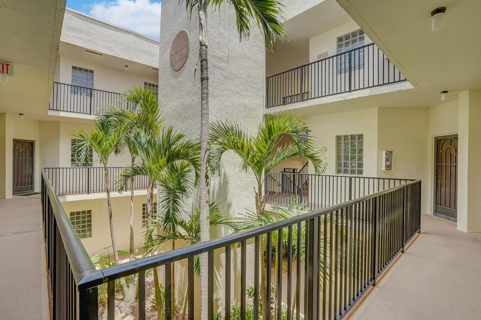 An open-air apartment complex courtyard features palm trees, walkways with black railings, and glass blocks — 5155 Europa Drive # I, Boynton Beach FL 33437, BOYNTON BEACH, FL