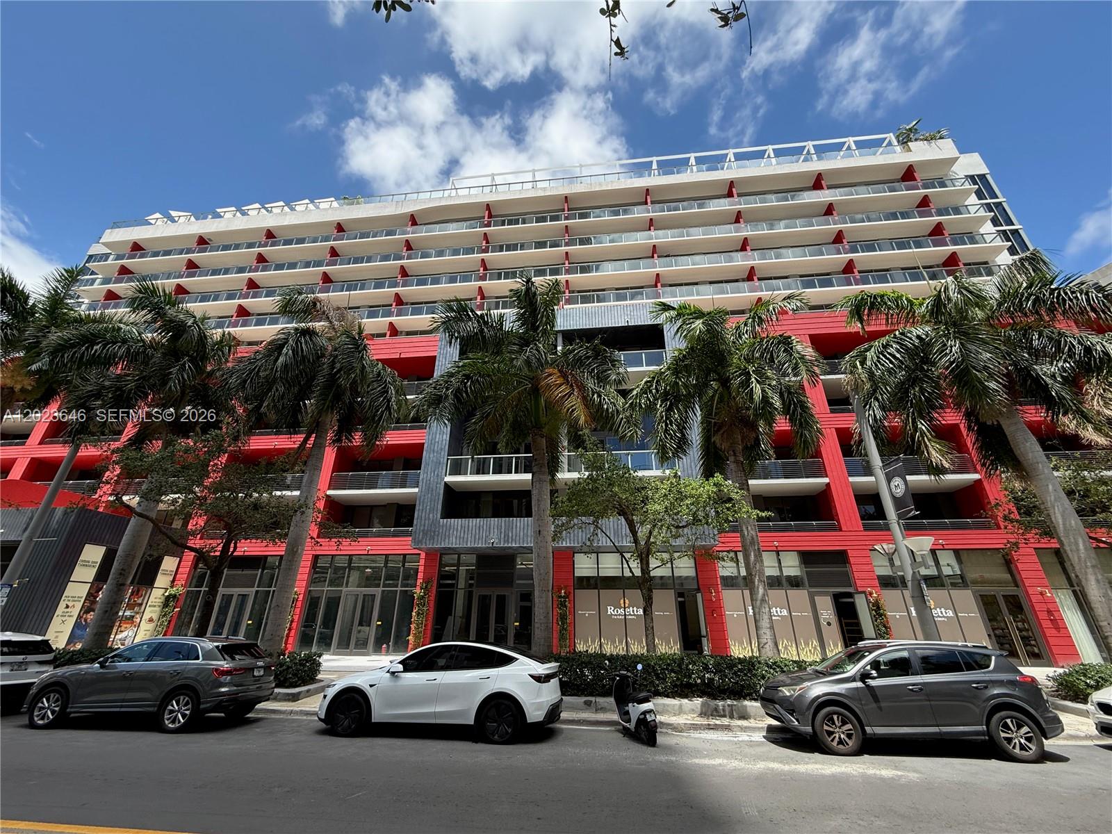A low-angle exterior view of a modern multi-story apartment building with red accents and palm trees — 90 NE 32 # 411, Miami FL 33137, MIAMI, FL
