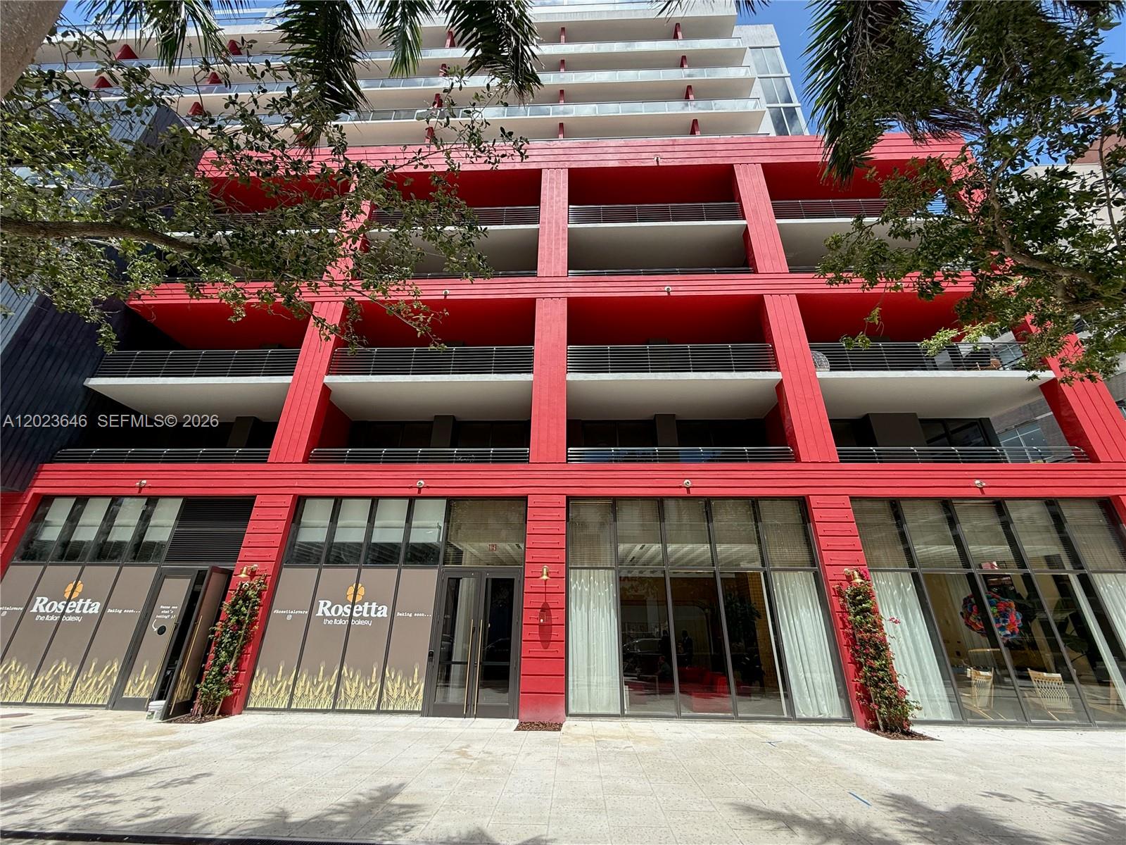 A low-angle exterior view of a modern building with red structural framing and storefronts — 90 NE 32 # 411, Miami FL 33137, MIAMI, FL