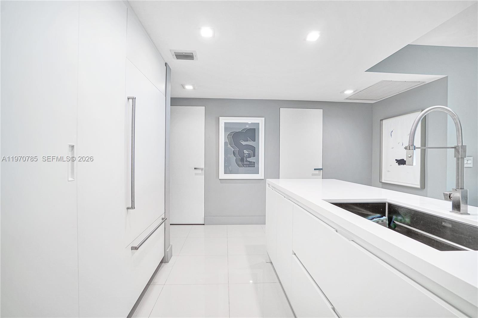 A modern, minimalist kitchen features white cabinetry, a central island with a sink, and artwork — 475 Brickell Ave # 1913, Miami FL 33131, MIAMI, FL