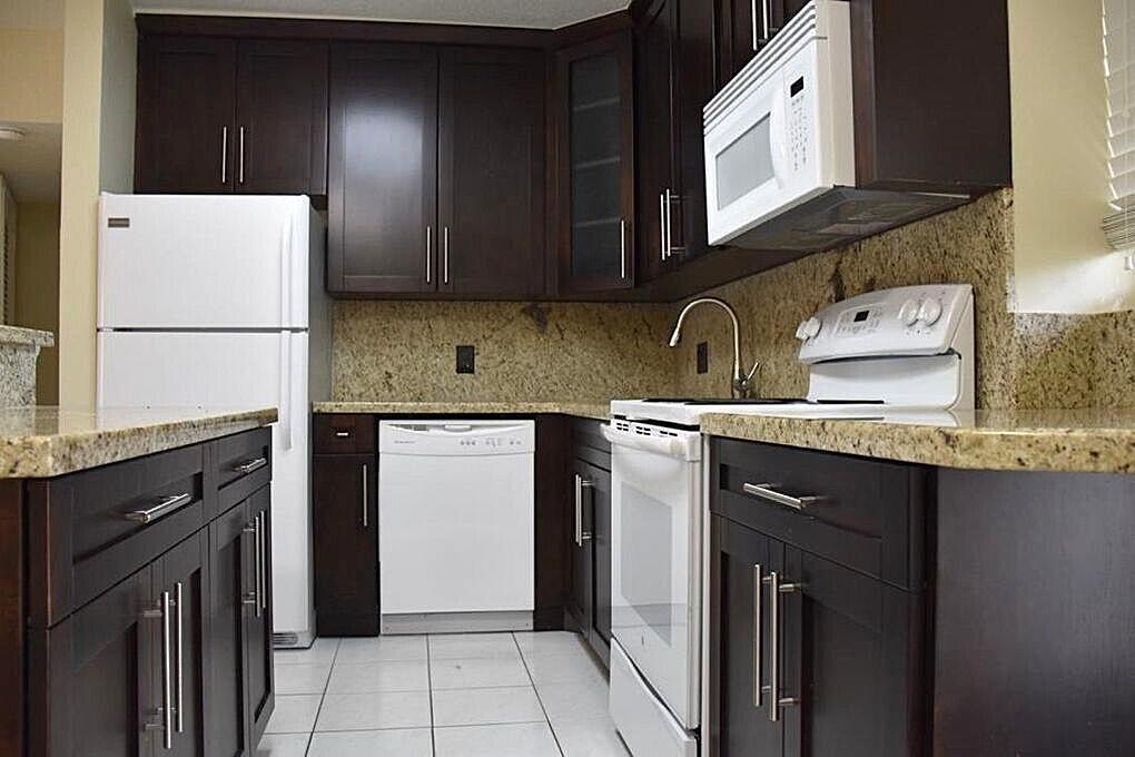 A kitchen features dark wood cabinets, granite countertops, white appliances, and light tile flooring — 1361 NE 27 Way, Pompano Beach FL 33062, POMPANO BEACH, FL