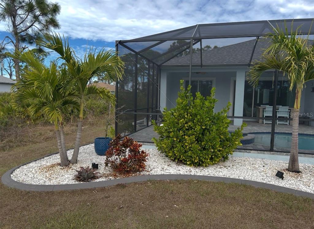 A landscaped garden bed with palm trees and shrubs sits beside a screened-in pool enclosure — 109 FLOWER ROAD, ROTONDA WEST, FL