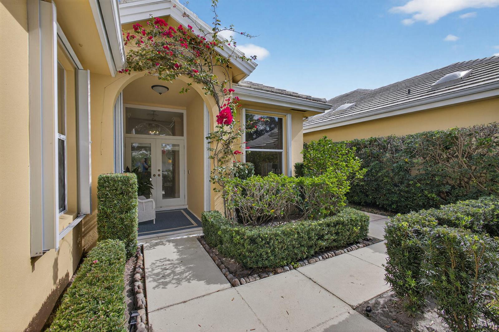 A yellow house exterior features a covered entryway, a concrete walkway, and manicured green hedges — 1180 Gator Trail, West Palm Beach FL 33409, WEST PALM BEACH, FL