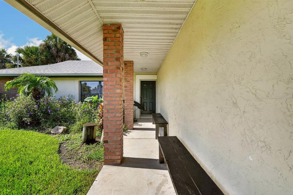A covered concrete porch features brick support columns, two wooden benches, and a dark front door — 109 CORAL REEF COURT N, PALM COAST, FL