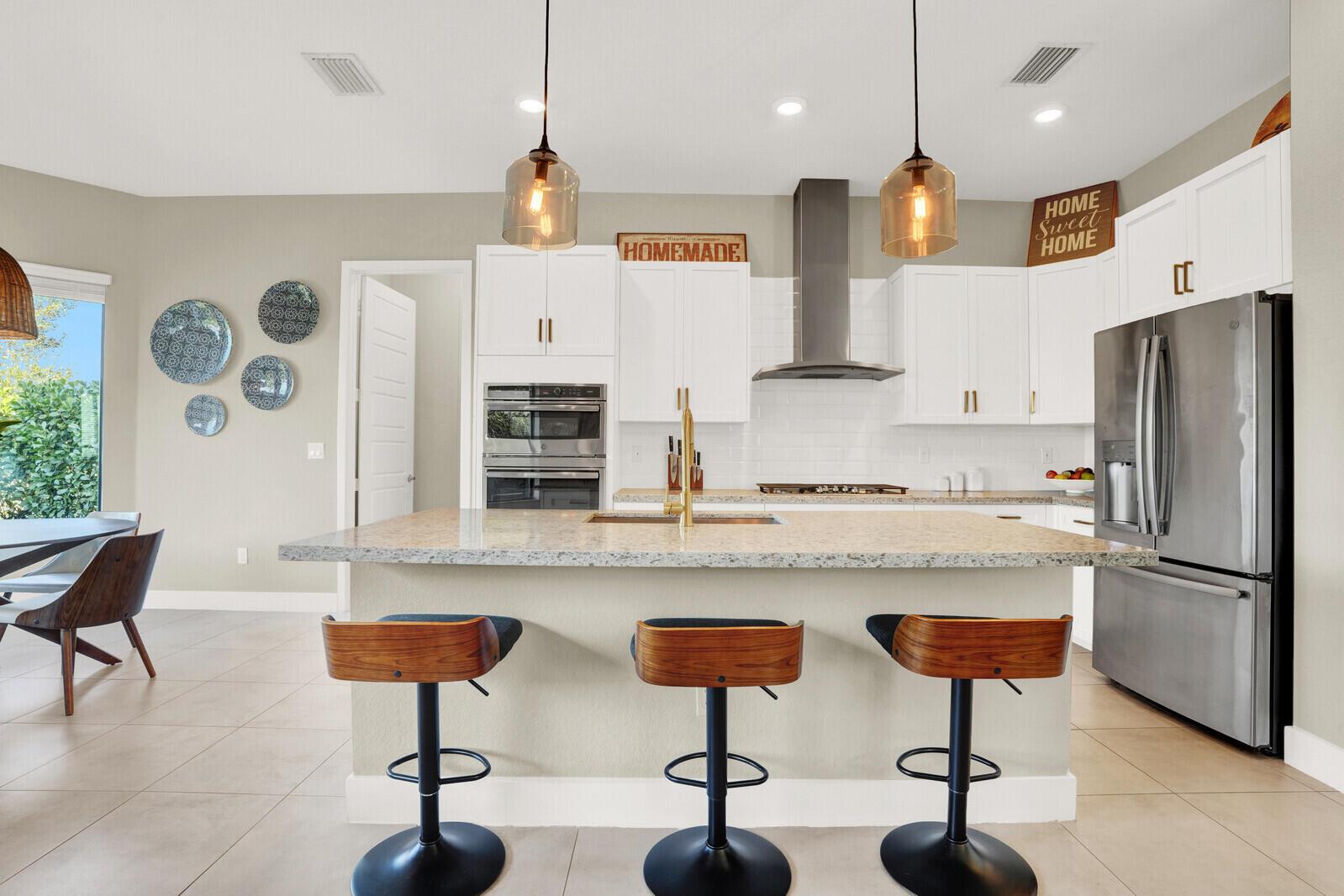 A kitchen features white cabinets, a granite island with three stools, and stainless steel appliances — 10767 Pacifica Way, Parkland FL 33076, PARKLAND, FL